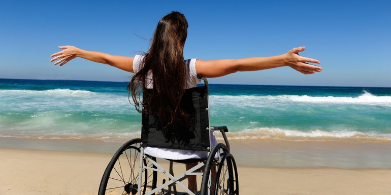 Photo from back of a girl sitting in a wheelchair infront of a beach with hands outstretched